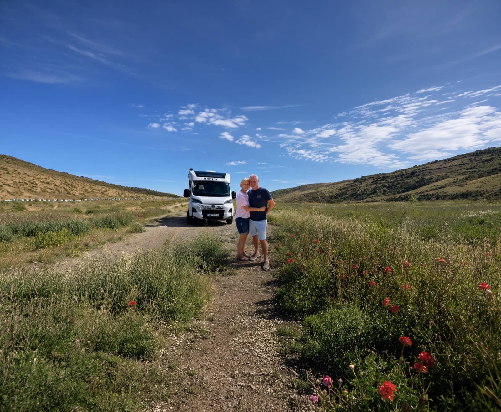 A couple standing beside their RV on a track through green rolling hills